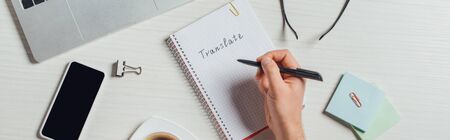 panoramic shot of translator writing in notepad on workplace with laptop and smartphoneの写真素材