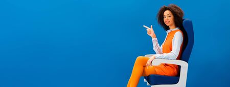 panoramic shot of smiling african american in retro dress sitting on seat and pointing with finger...