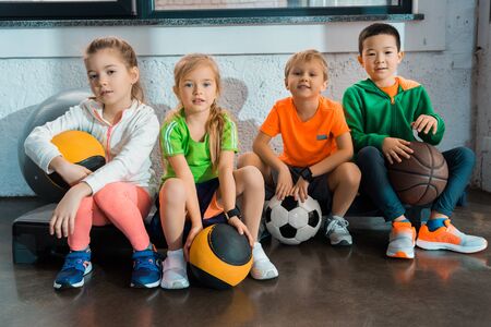 Multicultural children sitting on step platforms with balls in gymの写真素材
