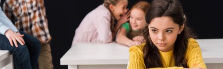 panoramic shot of upset schoolkid near classmates gossiping and laughing isolated on black, bullying conceptの写真素材