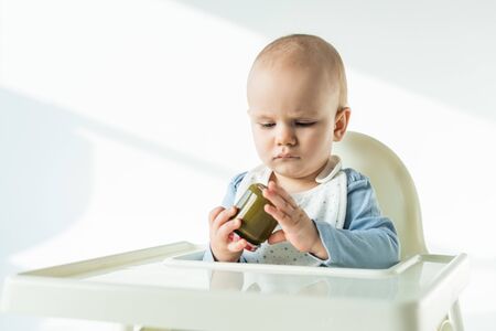Selective focus of cute baby holding jar of vegetable baby food on feeding chair on white background