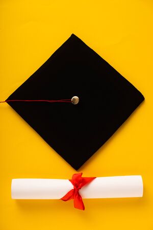 Top view of diploma with beautiful bow and black graduation cap on yellow backgroundの写真素材