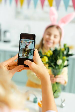 selective focus of mother taking photo of cute daughter in bunny ears with tulipsの写真素材