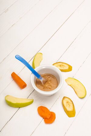 High angle view of baby food in bowl with spoon and sliced fruits and carrot on white wooden backgroundの写真素材