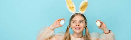 panoramic shot of cheerful kid with bunny ears holding painted easter eggs and looking away isolated...