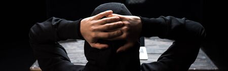 panoramic shot of hooded hacker with hands behind head isolated on black