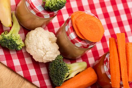 High angle view of jars of vegetable baby food with fresh vegetables and apple on tablecloth on wooden surfaceの写真素材