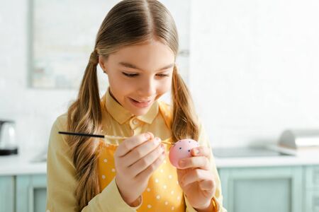 happy kid painting easter egg with paintbrush at home