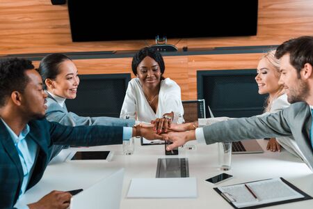happy, young multicultural businesspeople holding joined hands while sitting at desk in conference...