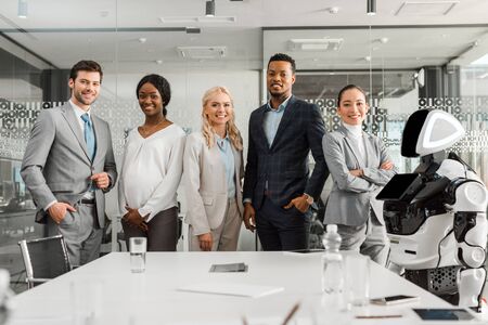 smiling multicultural businesspeople looking at camera while standing near robot in conference hall