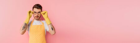 panoramic shot of confident young man in apron and rubber gloves touching glasses on pink background