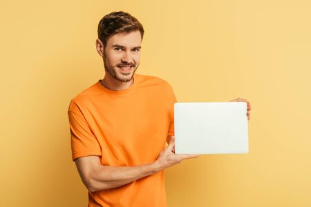 smiling young man showing closed laptop and looking at camera on yellow background