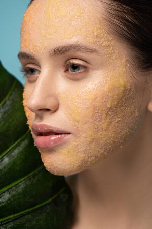 attractive girl with sugar scrub on face holding leaf, isolated on blue