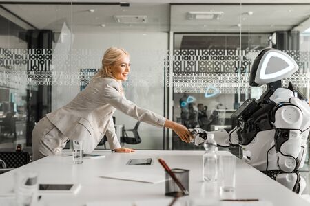 smiling businesswoman shaking hands with robot sitting at desk in meeting roomの写真素材
