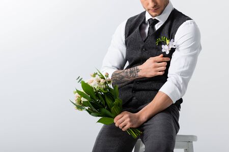 cropped view of tattooed elegant bridegroom with bouquet adjusting boutonniere on greyの写真素材
