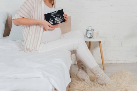 Cropped view of pregnant woman holding ultrasound scan of baby while sitting on bed