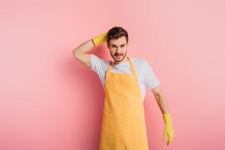 smiling young man in apron and rubber gloves touching head while looking at camera on pink backgroundの写真素材