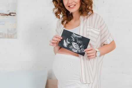 Cropped view of smiling pregnant woman showing ultrasound scan of baby in bedroomの写真素材