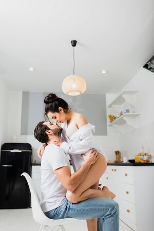 Side view of sexy girl in bra and shirt kissing boyfriend on chair in kitchenの写真素材