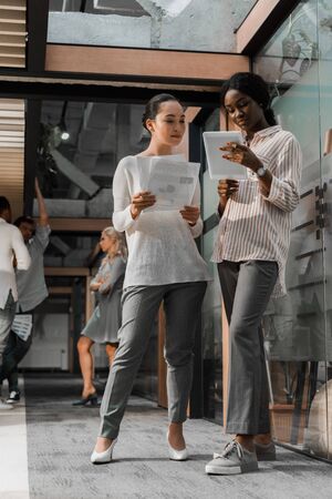 concentrated african american and asian businesswomen with digital tablet and documents standing in office with multicultural colleagues on backgroundの写真素材