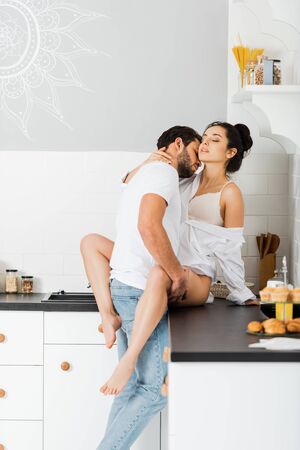 Handsome man kissing in neck and hugging seductive girlfriend in bra and shirt on kitchen worktopの写真素材