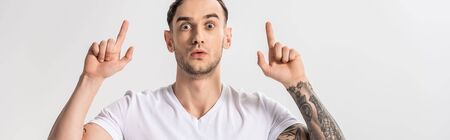 shocked handsome young tattooed man pointing with fingers up isolated on white, panoramic shot
