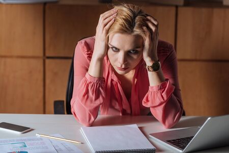 Frustrated woman holding head and sitting at table in officeの写真素材