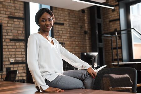 cheerful, pregnant african american businesswoman sitting on office desk and smiling at camera