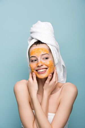 cheerful girl with citrus facial mask looking away and touching face isolated on blueの写真素材
