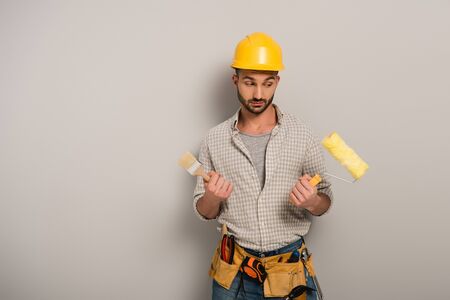 skeptical manual worker in hardhat holding paint roller and brush on grey