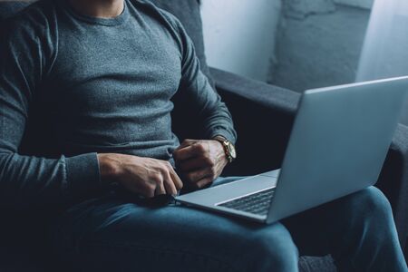 Cropped view of man unbuttoning jeans while watching pornography on laptop on sofaの写真素材