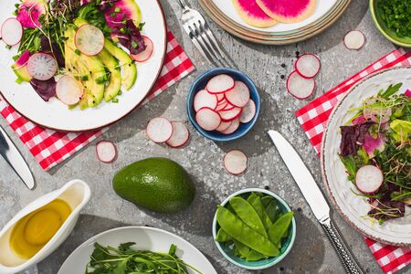 top view of fresh radish salad with greens and avocado served on plates on grey concrete surface with ingredients, cutlery and napkinsの写真素材