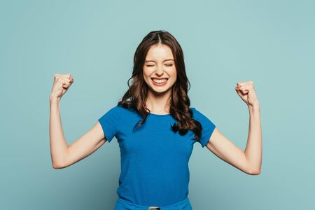 excited girl showing winner gesture with closed eyes while smiling on blue background