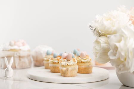 Selective focus of cupcakes on round board, decorative bunny and vase with flowers on white backgroundの写真素材
