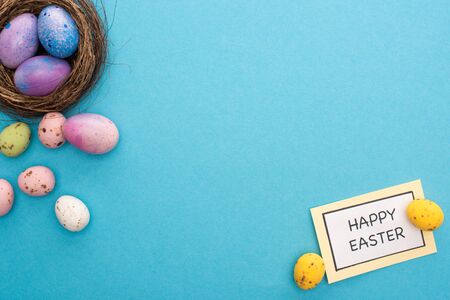 Top view of nest with colorful chicken and quail eggs with card with happy easter lettering on blue...