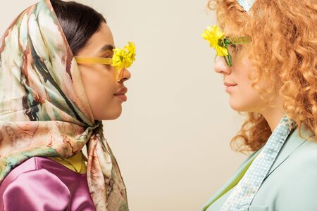 side view of african american girl and redhead woman in sunglasses with flowers looking at each other isolated on beigeの写真素材