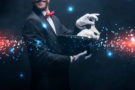 cropped view of smiling magician showing trick with white rabbit in hat, in dark room with smoke and...