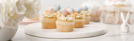 Selective focus of cupcakes on round board, decorative bunny and vase with flowers on white...