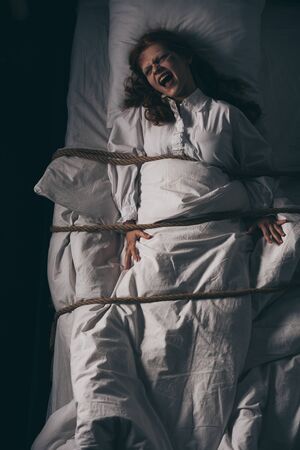 creepy yelling girl in nightgown bound with rope in bedの写真素材