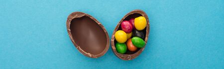 Top view of chocolate Easter egg halves with colorful sweets on blue background, panoramic shot