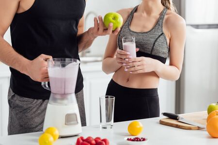 cropped view of man holding apple and blender near sportive girl with glass of smoothieの写真素材