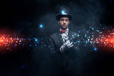 magician in suit and hat wearing gloves in dark smoky room with glowing illustrationの写真素材