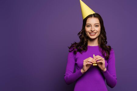 happy girl in party cap holding party horn while smiling at camera on purple background