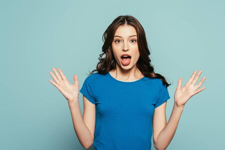 shocked girl looking at camera while standing with open arms isolated on blue