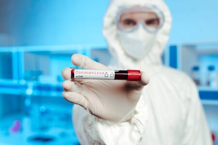selective focus of scientist in hazmat suit, medical mask and goggles holding sample with coronavirus letteringの写真素材
