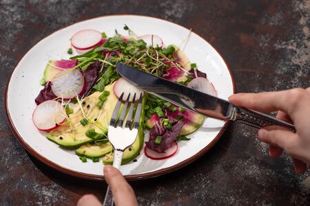 cropped view of woman eating fresh radish salad with greens and avocado with cutleryの写真素材