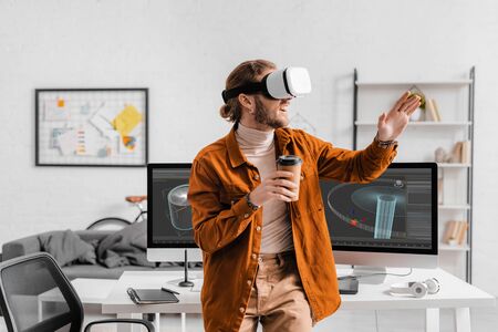 Cheerful digital designer in virtual reality headset holding coffee to go near 3d design project on computer monitors on tableの写真素材