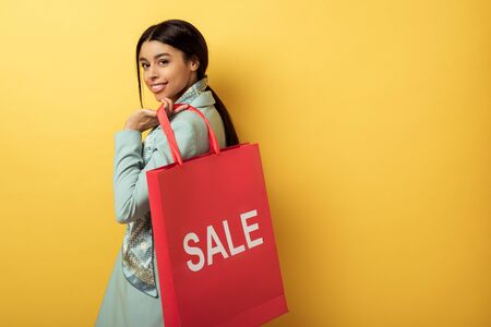 positive african american girl holding shopping bag with sale lettering and smiling on yellow