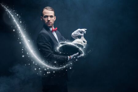 professional magician in suit showing trick with white rabbit in hat, dark room with smoke and...