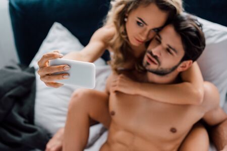 Selective focus of beautiful woman hugging muscular boyfriend while taking selfie with smartphone on bed isolated on greyの写真素材